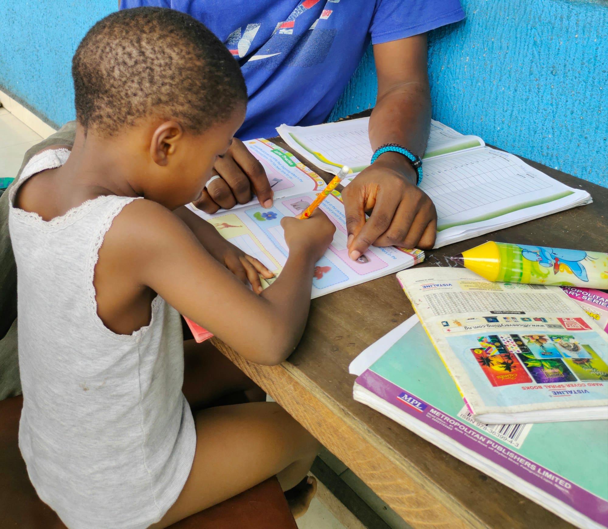 A young boy writes in an exercise book while an adult helps him