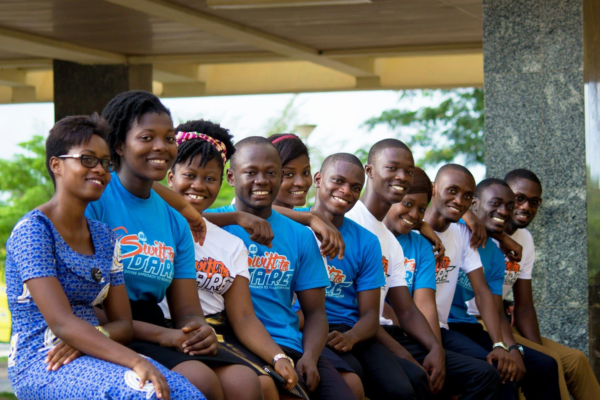 A row of smiling young women and men sitting together under a canopy